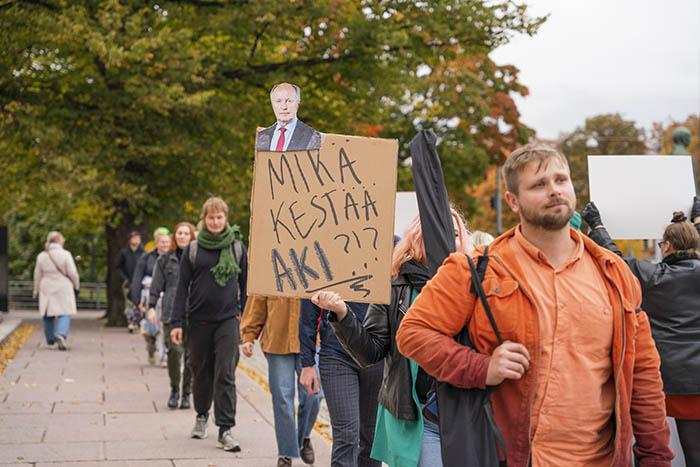 Kuvassa mielenosoittajia jonossa eduskuntatalon edessä. Kyltissä Aki Lindénin kuva ja teksti: "Mikä kestää Aki?"