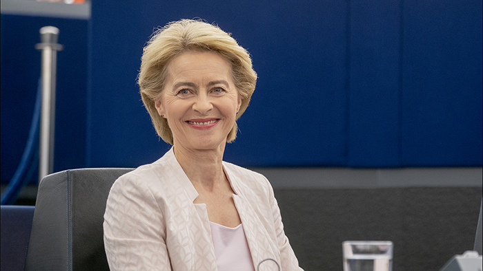 Kuvassa Euroopan komission nykyinen puheenjohtaja Ursula von der Leyen hymyilee ja katsoo kameraan. Hänellä on päällään vaalea jakku. Kuvan taustalla on tummansininen seinä.