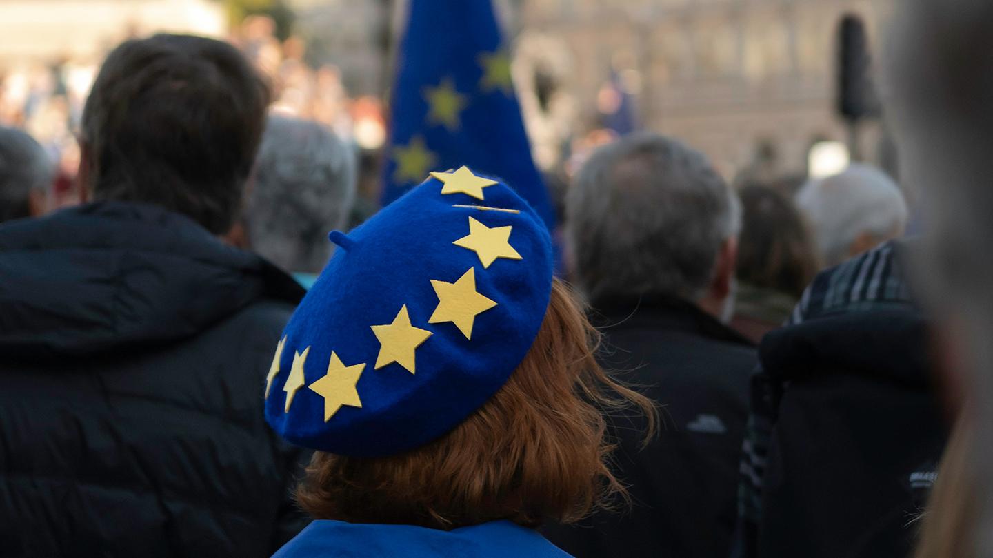 Nainen takaapäin kuvattuna kadulla ihmisjoukossa, päässään sininen, EU-tähdillä koristeltu baskeri.