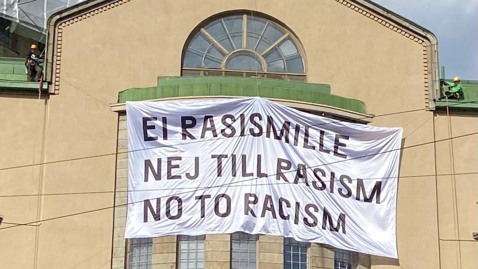 Ei rasismille -mielenosoituksen iso banderolli korkealla Uuden Ylioppilastalon yläikkunan edessä katolle kiipeilijöiden ripustamana.