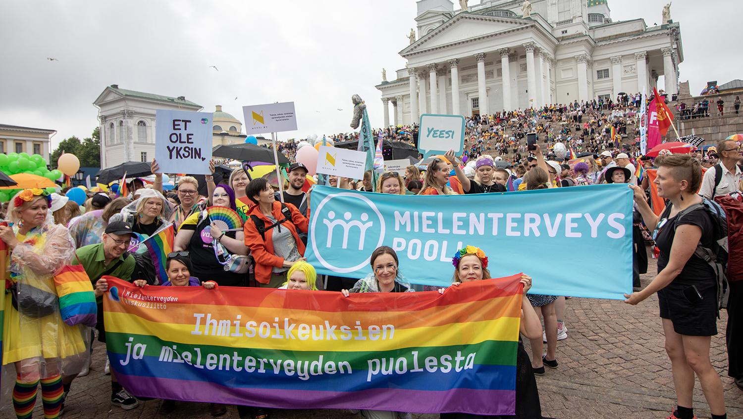 Pride-kokoontumisessa sateenkaaribanderolli ja mielenterveysbanderolli värikkäästi pukeutuneiden ihmisten kannateltavina Senaatintorilla Helsingissä.