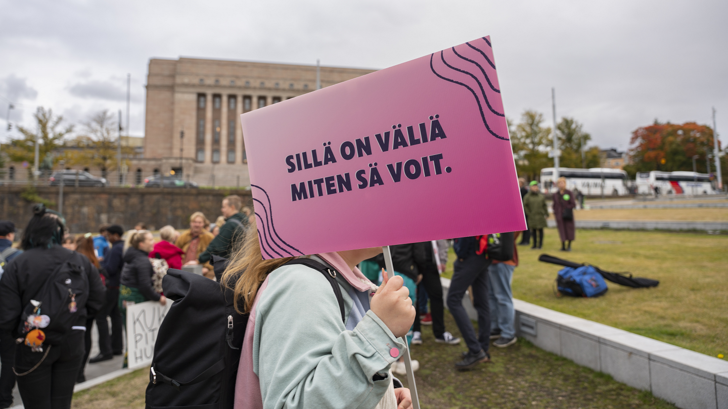 Mielenosoituksessa eduskuntatalon edustalla mielenosoittajan kasvot jäävät kyltin taakse. Kyltissä on teksti: Sillä on väliä miten sä voit.