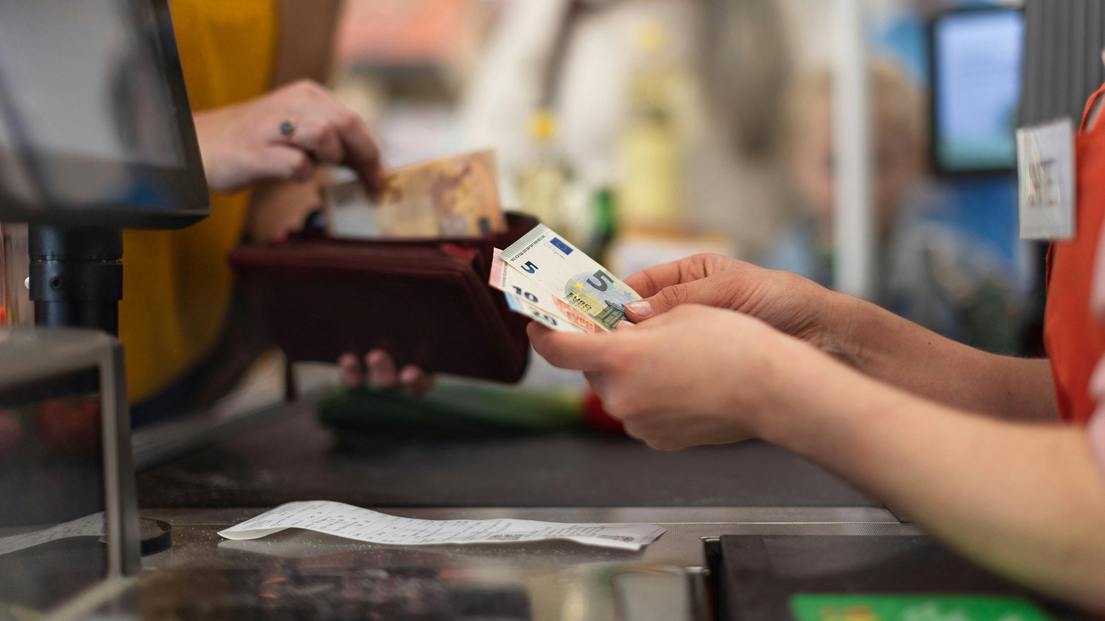 Kuvassa kassatyöntekijän seteleitä pitelevät kädet. Taustalla asiakkaan käsi nostaa seteliä avatusta lompakosta.