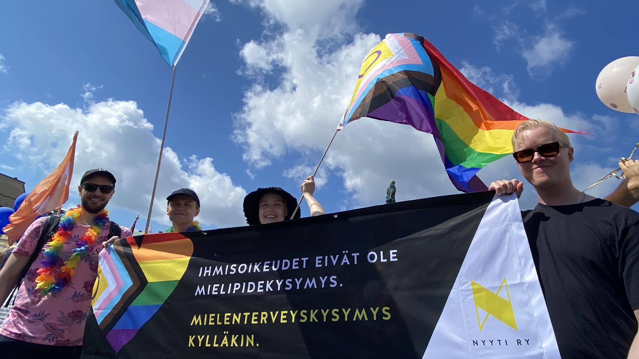 Liehuvien Pride-lippujen alla sinisen poutapilvitaivaan alla iloisesti hymyilevät ihmiset pitelevät banderollia jossa lukee: "Ihmisoikeudet eivät ole mielipidekysymys. Mielenterveyskysymys kylläkin."